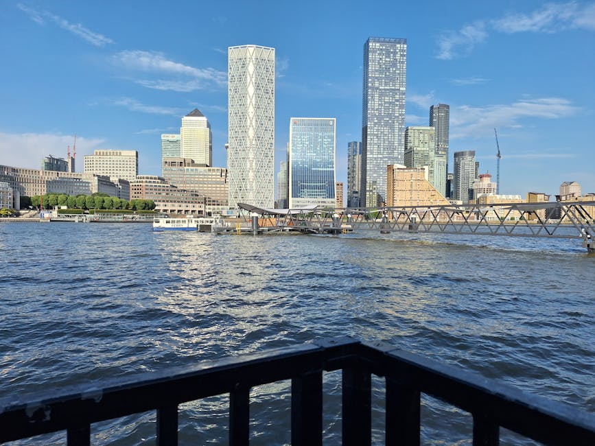 A view of the London city skyline taken from across the river, showing modern glass office buildings and skyscrapers under a clear blue sky. In the foreground, part of a black railing and a small sect
